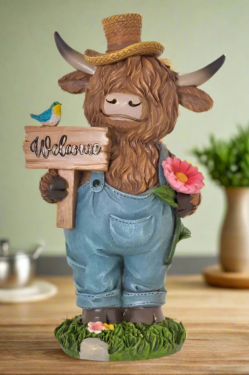 Garden figurine of a cow with a 'Welcome' sign, overalls, and a hat on a white background